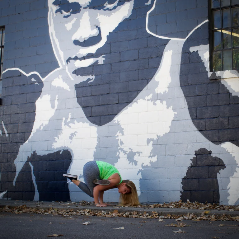 Ivy in crow pose on the pavement in front of a large painted portrait mural, autumn leaves around her