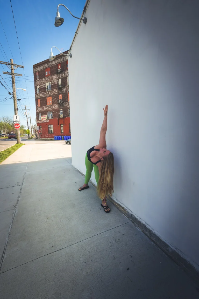 Ivy in a standing backbend with one hand on the studio's white side wall, the old brick Coal Hay Corn building across the street behind her