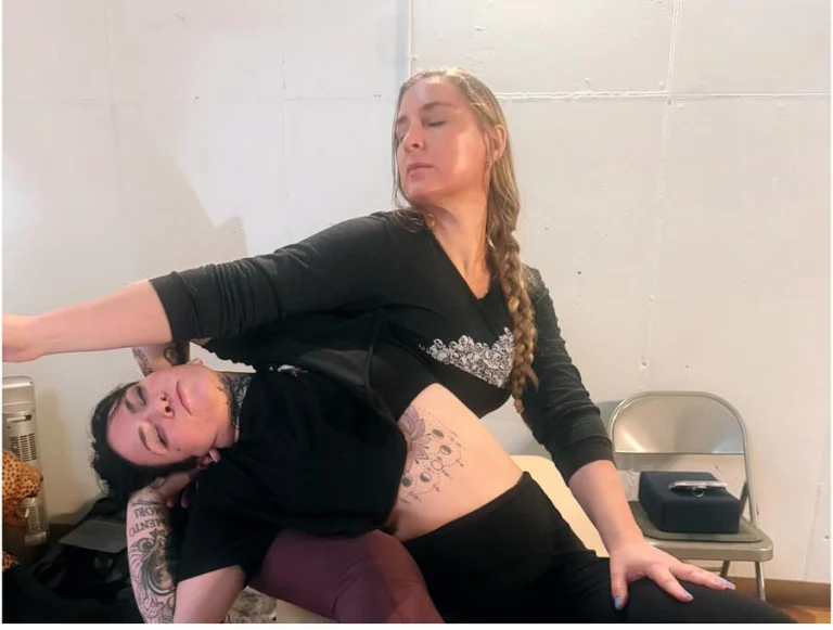 Ivy leaning a seated client into a side stretch against her own body, in the studio's white block room