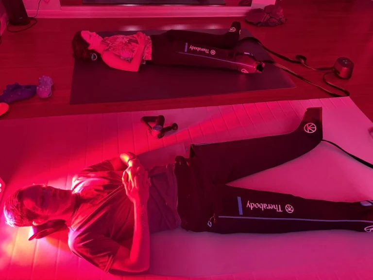 Two clients lying in compression boots under red light panels on mats side by side