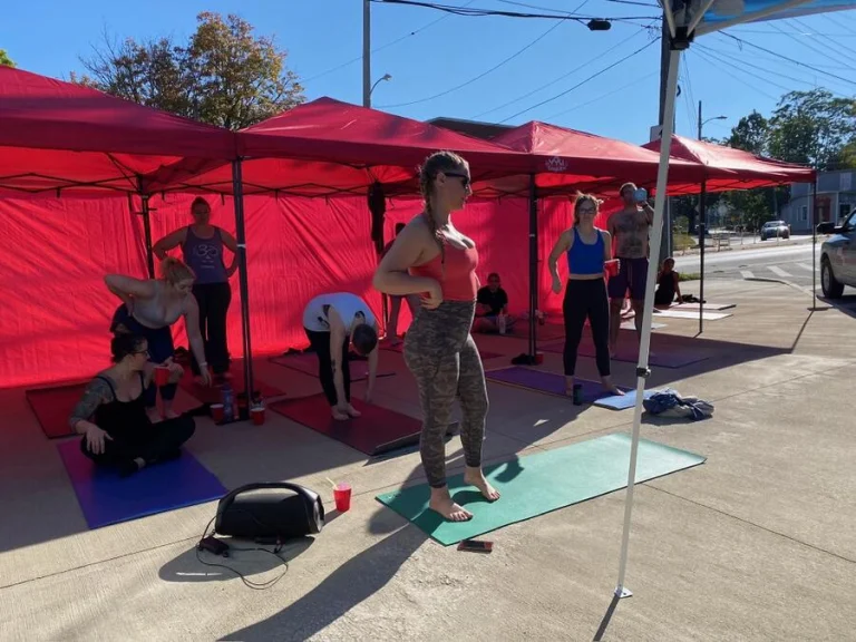 Students on mats under red canopies on the sidewalk outside the studio, one standing barefoot on a green mat