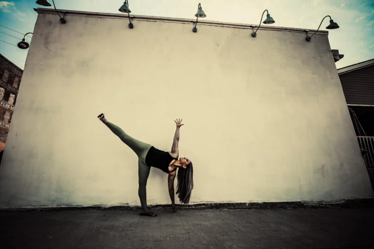 Ivy in half moon pose against a large blank cream wall under a row of gooseneck lights