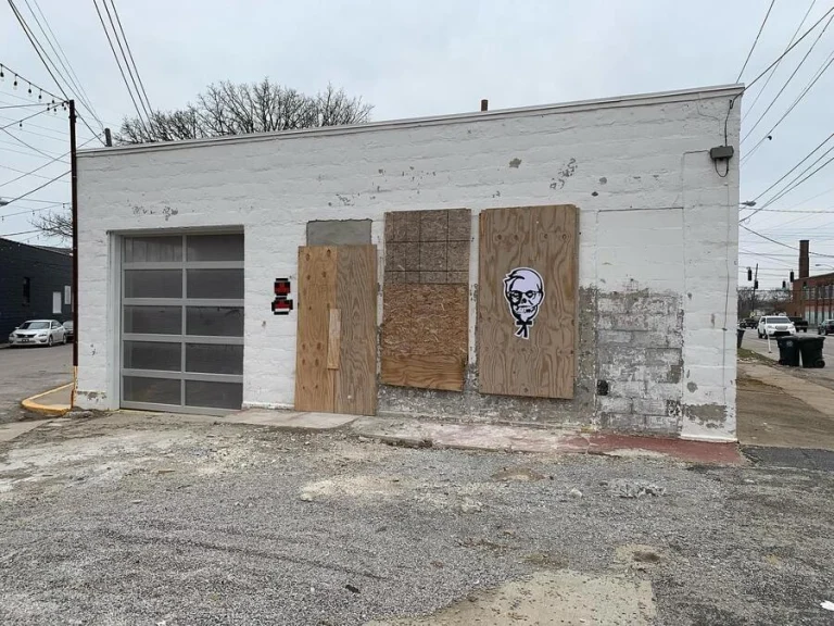 The boarded-up white block building the studio was built from, with graffiti on the plywood